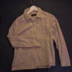 Beige overshirt från Riley - Säljer en beige overshirt från Riley i storlek S. Jackan har dragkedja framtill, tre fickor och klassisk krage. Tillverkad i mjuk bomull som känns chill och enkel att styla till olika outfits. Perfekt för lager på lager under vår och höst.
