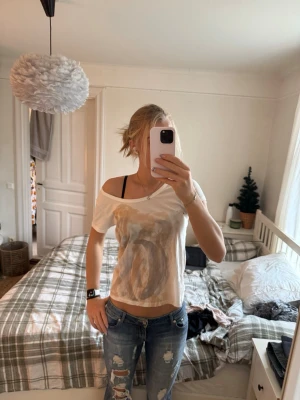 Vit croppad t-shirt från Lee - Säljer en vit croppad t-shirt från Lee med korta ärmar och ett stort abstrakt tryck i beige och bruna toner framtill. T-shirten har en avslappnad passform och en offshoulder-stil som ger en cool vibe. Tillverkad i mjuk bomull. Aldrig använd utan prislapp. 