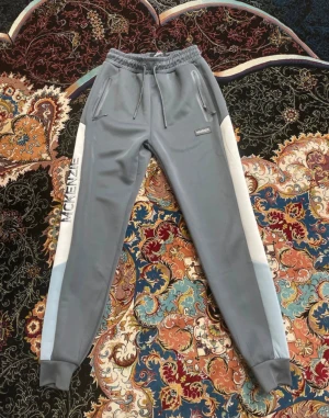 mjukisbyxor från McKenzie - Snygga grå mjukisbyxor från McKenzie med vita sidopaneler och logotyp längs benet. Byxorna har snörning i midjan, dragkedjeförsedda fickor och ribbade muddar vid bensluten. Perfekta för chill eller träning, tillverkade i mjukt syntetmaterial.