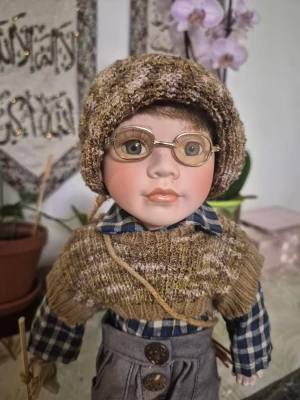 Vintage GEPPEDDO Porslin Vit Bondepojke Blå Ögon m. Glasögon Docka Storlek 45 cm - En charmig porslinsdocka med detaljerade kläder och glasögon. Dockan har en mössa, en rutig skjorta, en stickad väst och grå byxor med lappar. Den håller i en liten kvast och har en liten bunt med pinnar på ryggen.