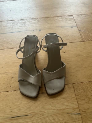 Beige sandaler med klack från Rasgini - Säljer ett par stilrena beige sandaler från Rasgini med låg klack och smala remmar. Skorna har fyrkantig tå och en snygg, enkel design som passar till många olika outfits. Materialet är syntet och färgen är ljusbeige. Perfekt för dig som gillar minimalistisk stil.