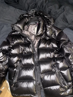 Svart pufferjacka från Moncler - Säljer en svart pufferjacka från Moncler med glansig finish och huva. Jackan har dragkedja framtill och två sidofickor med dragkedja. Insidan har ett unikt serietidningsmönster från Moncler. Perfekt för kalla dagar och riktigt snygg stil.