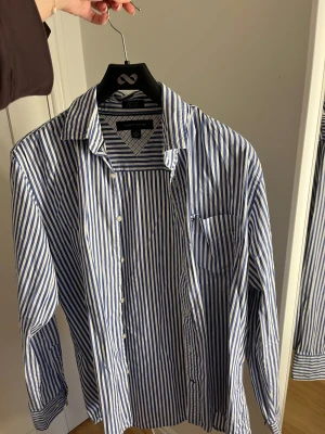 Blåvit randig skjorta Tommy Hilfiger - Snygg blåvit skjorta från Tommy Hilfiger i klassisk randigt mönster. Skjortan har lång ärm, bröstficka med liten logga och vita knappar framtill. 