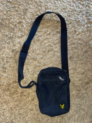 Svart axelväska från Lyle & Scott - Svart axelväska från Lyle & Scott med justerbar rem och gul logga framtill. Väskan har två dragkedjor upptill och ett extra fack med dragkedja på framsidan. Tillverkad i slitstark polyester och har en kompakt, rektangulär form. Lite sliten på baksidan som man ser på bild 3.