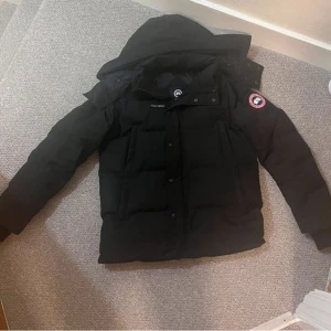 Svart dunjacka från Canada Goose - Säljer en svart dunjacka från Canada Goose med huva och klassisk logga på ärmen. Jackan har en puffer-design, flera fickor framtill och stängs med både dragkedja och knappar. Perfekt för kalla dagar och har en riktigt clean look.