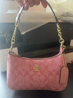 Coach axelväska rosa  - Säljer en snygg rosa axelväska från Coach med deras klassiska logomönster. Väskan har guldfärgade kedjedetaljer, dragkedja och en avtagbar axelrem. Materialet är tålig polyester och väskan har en stilren, kompakt form som är perfekt för att bära med sig det viktigaste.