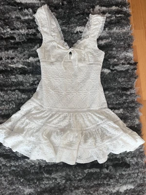 Vit spetsklänning från Gina Tricot (S/M) - Supersöt vit kortklänning från Gina Tricot i storlek S. Klänningen har spetsdetaljer, volanger och ett knyt framtill vid urringningen. Materialet är bomull med broderade mönster och spets längs axelbanden. Perfekt för sommarens alla tillfällen!