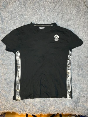 Svartvit Urban Street t-shirt  - Svart t-shirt från Terranova med oversized passform och tryck 'Urban Street' på bröstet. Snygga vita band med text och rutmönster längs sidorna ger en cool streetkänsla. Tillverkad i mjuk bomull, perfekt för dig som gillar avslappnad stil.