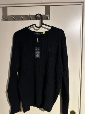 Svart kabelstickad tröja Polo Ralph Lauren - Svart kabelstickad tröja från Polo Ralph Lauren i storlek M. Klassisk rund hals och långärmad modell med röd broderad logga på bröstet. Tröjan är gjord i mjuk ull och har ribbade muddar vid ärmslut och nederkant. Perfekt för dig som gillar stilrena och tidlösa plagg.