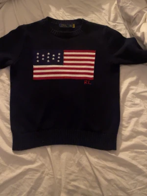 Mörkblå stickad tröja Polo Ralph Lauren - Snygg mörkblå stickad tröja från Polo Ralph Lauren med amerikansk flagga i rött, vitt och blått på bröstet. RL-broderi i rött nedtill på flaggan. Rund hals och långa ärmar, perfekt för dig som gillar klassisk college-stil.