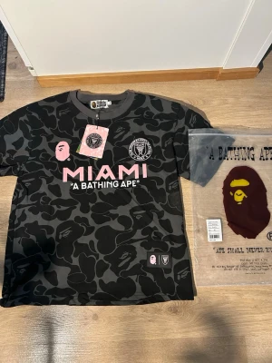 Svart camo BAPE x Inter Miami t-shirt - Säljer en limited edition t-shirt från BAPE x Inter Miami med svart och grått camo-mönster, rosa tryck och patchar. Stor MIAMI-logga fram och BAPE 93 på ryggen. Kortärmad med rund hals och coola detaljer. Perfekt för dig som gillar streetwear och fotboll.