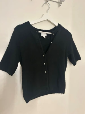 Svart stickad kofta från H&M - Svart stickad kofta från H&M i storlek XXS. Koftan har korta ärmar, rund halsringning och knäpps med silvriga knappar framtill. Mjuk och enkel design som funkar till många olika stilar. Perfekt för lager-på-lager under kyliga dagar.