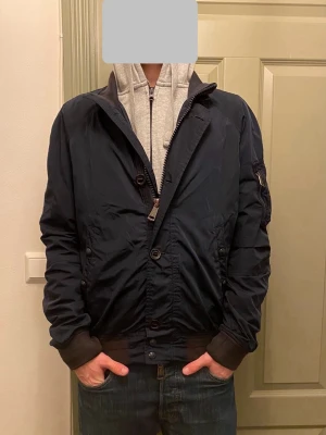 Mörkblå bomberjacka från Polo Ralph Lauren  - Säljer en riktigt snygg mörkblå bomberjacka från Polo Ralph Lauren, med bruna ribbade muddar och krage. Jackan har både dragkedja och knappar framtill samt en ficka med dragkedja på ärmen. Liten defekt (kolla bild 5) men int esp synligt. Vid funderingar, fråga på! 