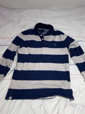 Randig långärmad henley från Gant - Snygg långärmad henley från Gant med breda marinblå och ljusgrå ränder. Skjortan har klassisk krage, knappar vid halsen och är tillverkad i mjuk bomull. Perfekt för en avslappnad och stilren look.