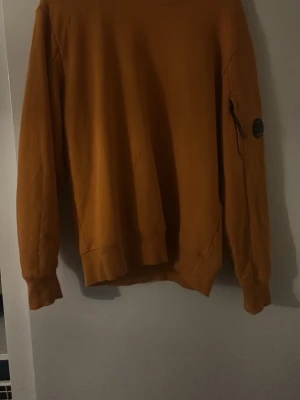 Orange sweatshirt från C.P. Company - Säljer en orange sweatshirt från C.P. Company med klassisk rund hals och långärmad design. Tröjan har en ikonisk linsdetalj på ärmen och är tillverkad i mjuk bomull. Använd väldigt få gånger så mycket fin