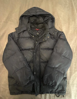 Svart barn pufferjacka från GANT  - Svart barn pufferjacka från GANT i storlek 158/164cm . Jackan är tillverkad i nylon och har dunfyllning för extra värme. Klassisk design med elastiska muddar och knappar vid kragen. Perfekt för kalla dagar.