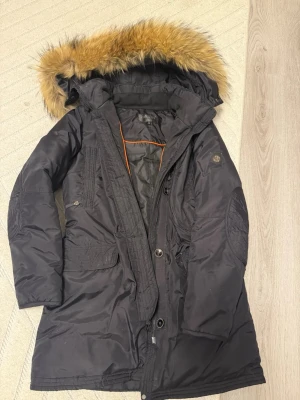 Svart parkajacka med päls från Hollies - Parkajacka från Hollies i svart med snygg fuskpäls på huvan och orange detaljer på insidan. Jackan har långa ärmar, flera fickor och stängs med både dragkedja och knappar. Perfekt för kalla dagar och har en klassisk, stilren look.