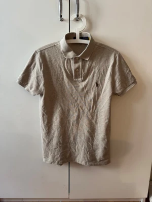 Beige pikétröja från Polo Ralph Lauren - Säljer en beige pikétröja från Polo Ralph Lauren i slim fit-modell. Tröjan har klassisk krage, korta ärmar och knäppning med två knappar framtill. Materialet är mjuk bomull och den har den ikoniska broderade loggan på bröstet.