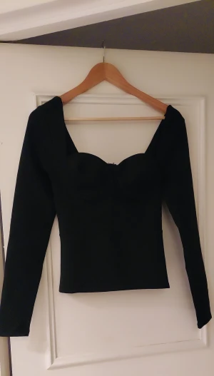 Svart långärmad korsett-topp från Nelly  - Säljer en svart korsett-topp med lång ärm och markerade kupor. Toppen har en snygg, figurnära passform och en stilren, minimalistisk look. Materialet är slätt och stretchigt, perfekt för att ge en cool siluett. Dragkedja framtill för enkel påklädning.