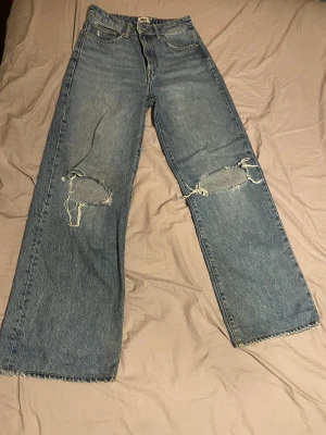 Blå wide jeans med hål i  knän - Säljer ett par blå jeans från laget 157 med breda ben och råa, slitna hål på båda knäna. Modellen är high waist och har klassiska fem fickor, bälteshällor och dragkedja. Perfekt för dig som gillar en avslappnad och edgy stil.