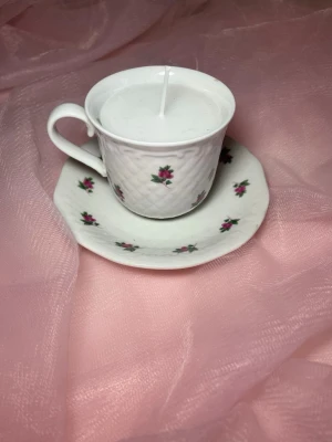 Doftljus i en tekopp i en vit färg med rosa blommor på  - Det här ljuset är gjort av återanvändbara material. Tekoppen är köpt secondhand och stearinet är från kyrkor som tänkte slänga det. Det här ljuset har en svag doft utav vanilj, så känsliga även kan använda det.