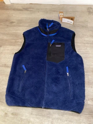 Blå fleeceväst från Patagonia M - Mörkblå fleeceväst från Patagonia med hög krage och tre fickor med dragkedja. Västen har en svart bröstficka och blå detaljer vid dragkedjorna. Perfekt för lager-på-lager och outdoor. Materialet är mjuk fleece som håller värmen.