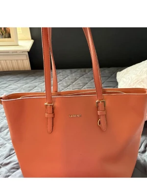 Rosa axelväska från Vabene - Snygg rosa axelväska från Vabene med stilrena guldfärgade detaljer och praktisk dragkedja upptill. Väskan har två långa axelremmar och en enkel, klassisk design som passar perfekt för dig som vill ha en rymlig och trendig väska.