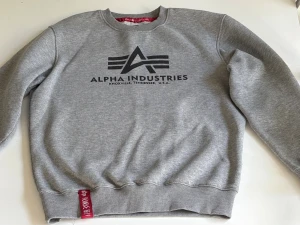 Grå sweatshirt från Alpha Industries - Snygg grå sweatshirt från Alpha Industries med svart logotryck på bröstet och en röd detalj vid nederkanten. Tröjan har rund hals, ribbade muddar och är tillverkad i mjuk bomullsmix. Perfekt för chill dagar och streetwear-stil.