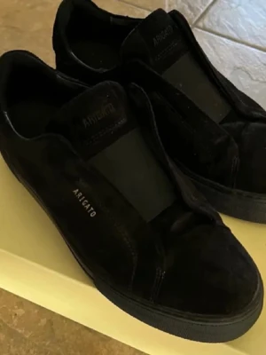 Svarta sneakers från Axel Arigato - Säljer ett par svarta sneakers från Axel Arigato med stilren design och diskret logga på sidan. Skorna har rund tå och platt sula, tillverkade i mocka för en lyxig känsla. Perfekta för dig som gillar minimalistisk stil.