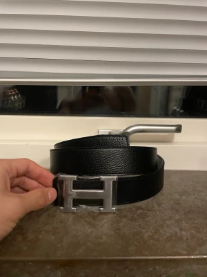 Bälte med H-spänne - Bilden visar ett svart bälte med en stor H-formad metallspänne, inte en väst. Ingen information om väst kan ges. Den kan bli till vit 