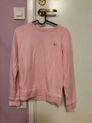 Ljusrosa sweatshirt från GANT - Säljer en ljusrosa sweatshirt från GANT med rund hals och diskret logga på bröstet. Tröjan har långa ärmar och ribbade muddar vid ärmslut och nederkant. Perfekt för dig som gillar stilrena och bekväma plagg.