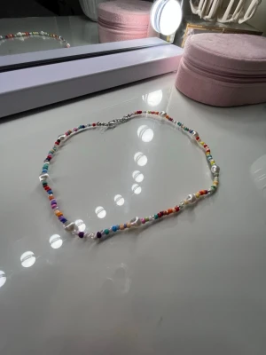 Färgglad pärlhalsband med vita inslag - Unikt halsband med små färgglada pärlor och vita pärlimitationer. Halsbandet har en enkel, smal design och stängs med en metallhake. Perfekt för dig som vill sticka ut med accessoarer i regnbågens alla färger.