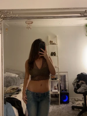 Brun spetsväst med v-ringning - Säljer en brun croppad väst i spets med v-ringad hals och fina spetsdetaljer längs kanterna. Västen är figurnära och har en öppen rygg som ger en snygg och trendig look. Perfekt att styla över ett linne eller som den är.