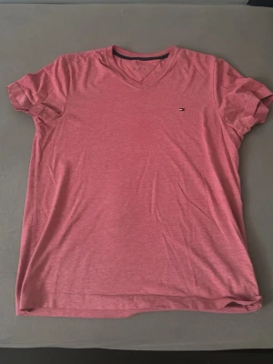 Rosa v-ringad t-shirt Tommy Hilfiger - Säljer en rosa t-shirt från Tommy Hilfiger med v-ringning och liten logga på bröstet. T-shirten är i mjuk bomull och har korta ärmar. Perfekt basic-plagg med snygg passform och diskret branding.