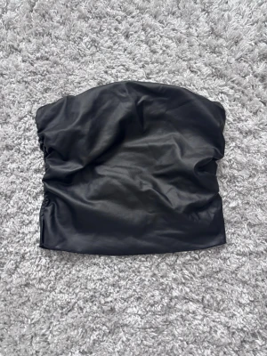 Svart tubtopp  - Säljer en svart tubtopp från SHEIN i storlek XS. Toppen är gjord i fejkskinn (polyester) och har snygga rynkade detaljer på båda sidorna som ger en cool look. Perfekt att styla med höga jeans eller kjol för en trendig outfit.