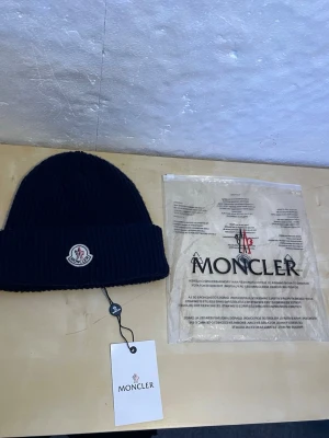Marinblå Moncler mössa i ull - Säljer en marinblå ribbstickad mössa från Moncler med klassisk logotyp framtill. Mössan är tillverkad i mjuk ull och har en vikt kant för extra stilpoäng. Perfekt accessoar för kyliga dagar och snygg till alla outfits.