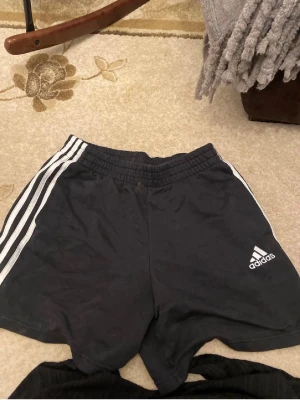 Svarta Adidas shorts med vita ränder - Klassiska svarta Adidas shorts med vita ränder längs sidorna och Adidas-logga på ena benet. Elastisk midja för skön passform. Perfekta för träning eller chill. Tillverkade i mjuk bomullsmix.