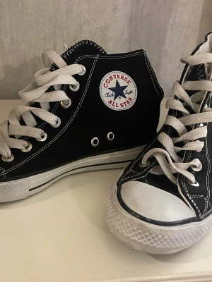 Svarta Converse All Star high-tops - Klassiska svarta Converse All Star high-tops med vit tåhätta, vita snören och kontrasterande vita sömmar. Skorna har rund tå, platt sula och ikonisk logga på sidan. Tillverkade i slitstark bomullscanvas med gummisula. Perfekta för en avslappnad och cool stil.