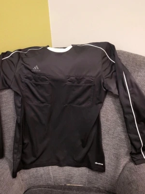 Adidas Referee 16 long sleeve svart tröja, storlek M - Svart långärmad Adidas tröja i funktionsmaterial med vita detaljer och två bröstfickor. Tröjan har rundad halsringning med vit kant och diskret Adidas-logga på bröstet. Tillverkad i ventilerande climacool-polyester, perfekt för sport och aktivitet.