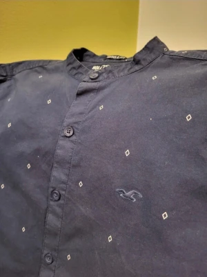 Mörkblå Hollister henley kortärmad S - Kortärmad mörkblå henley från Hollister med diskret diamantmönster och broderad logga på bröstet. Skjortan har muscle fit-passform, rund krage utan slag och knappar framtill. Tillverkad i mjuk bomull med lite stretch för extra komfort.