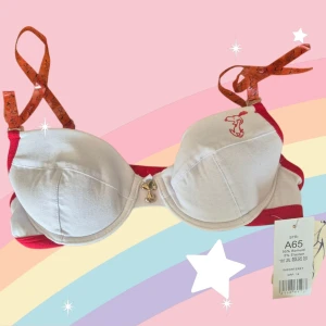 Snoopy BH 65A - Söt vit Snobben eller Snoopy-behå i storlek 65A. Avtagbara halvgenomskinliga orange'a axelband. Uttagbara kuddar. Fin berlock mellan kuporna. Varan är oanvänd och kommer från ett konkurslager från 2000-talet. I många fall är prislappen kvar (se bild). Har fler annonser och bra bundlerabatt vid köp av flera.