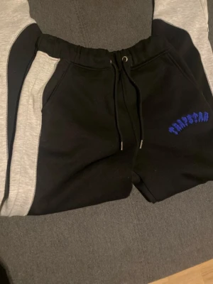 Trapstar mjukisbyxor svart/grå S - Trapstar mjukisbyxor i svart med gråa paneler längs benen och blå Trapstar-logga broderad på ena sidan. Byxorna har dragsko i midjan och är tillverkade i mjukt material för maximal komfort. Perfekta för chill eller streetwear.