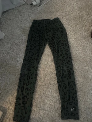 Leopardmönstrade leggings från SOC - Snygga gröna leggings från SOC med svart leopardmönster. De har en tight passform och är tillverkade i ett stretchigt syntetmaterial som passar perfekt till träning eller chill. Midjan är normal och tyget är mjukt och följsamt. Strl 146/152.