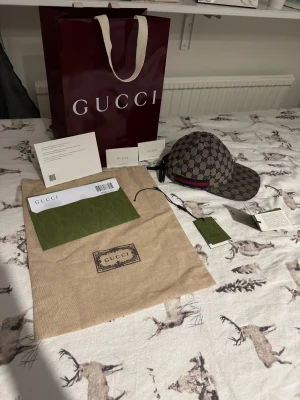 Gucci keps i XS - Snygg Gucci keps i klassisk GG grå och mörkblå färg. Kepsen har en böjd skärm, justerbar rem bak och en ikonisk röd-blå rand på sidan. Materialet är slitstark canvas med bomullsfoder. Perfekt för dig som vill ha en lyxig streetstyle-look. Kvitto, påse, dustbag, taggs, tack kort allt finns. Storlek XS