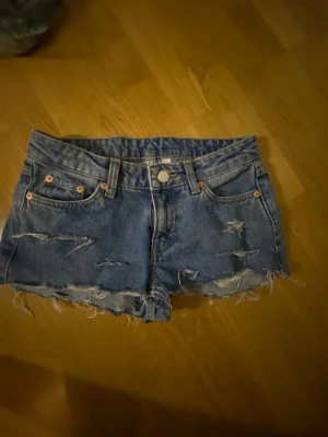 super låg midjade blå hål jeans shorts  - Säljer ett par blå jeansshorts med råa, fransiga kanter och slitna detaljer framtill. Klassisk femficksmodell med knapp och dragkedja. Perfekta för sommaren och ger en avslappnad vibe till din outfit.