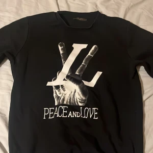 Svart Louis Vuitton sweatshirt Peace & Love - Säljer en svart sweatshirt från Louis Vuitton med stort vitt tryck av en hand som gör peace-tecken och texten 'PEACE AND LOVE' framtill. Tröjan har rund hals och långa ärmar. Perfekt statement-plagg för dig som gillar streetstyle.