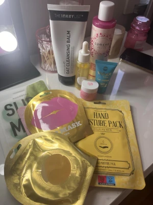 Hudvårds- och sminkkit med sheet masks - Ett kit med flera sheet masks från Kocostar, inklusive hand moisture pack och lip mask i guld och rosa förpackningar. Inkluderar även The Inkey List Oat Cleansing Balm, Lumene ansiktsvatten, samt små tuber och burkar med serum och läppbalsam. Perfekt för en self-care kväll.