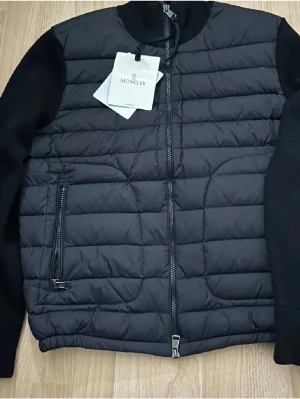 Marinblå cardigan från Moncler - Säljer en marinblå cardigan från Moncler med quiltad front och stickade ärmar. Jackan har dragkedja framtill, ribbad krage och muddar samt Moncler-logga på ärmen. Perfekt för dig som vill ha en stilren och exklusiv look.