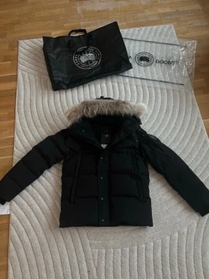 Canada Goose Wyndham Black Label  - Säljer en Canada Goose Wyndham Black Label  dunjacka med äkta päls på huvan. Jackan har klassisk patch på ärmen, dragkedja och knappar framtill samt stora fickor. Perfekt för kalla vinterdagar och riktigt snygg passform.