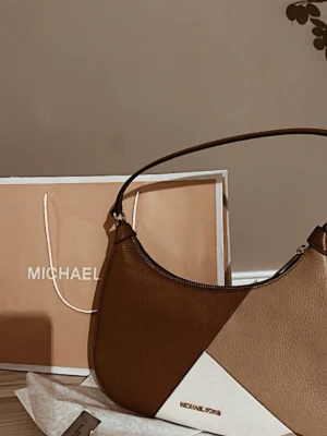 Michael Kors axelväska  - Säljer en snygg axelväska från Michael Kors i brunt, beige och vitt skinn. Väskan har en modern bågformad design och guldfärgad logga framtill. Perfekt storlek för det viktigaste och enkel att bära över axeln.